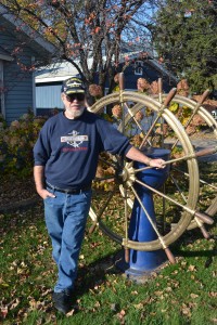 Capt. Tom Mackay, a retired captain from the Vista Fleet, stands with a big lake freighter wheel in the front yard of his home on Park Point. Photo by Jenae Peterson.