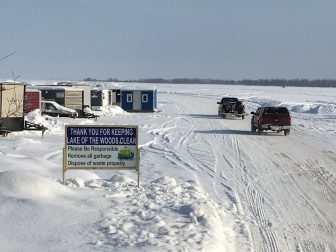A snowy, frozen lake with a sign in the foreground and cars and buildings in the background. 